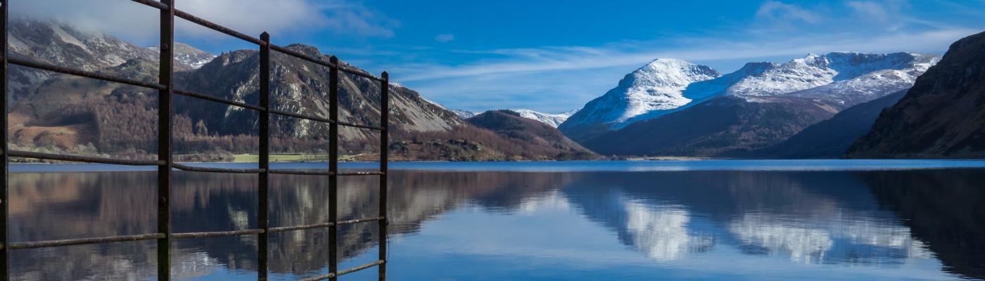 Ennerdale Water taken from the level of the shoreline. Railings descending into the water from the left are visible in the foreground. In the background are snow-capped mountains.