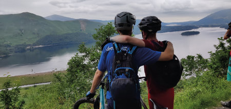Couple of mountain bikers with arms around one another looking out at scenery including a lake