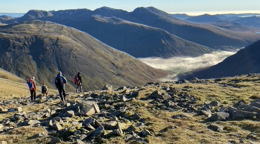 A view from the summit of Pillar looking out over surrounding dark green hilltops. Rocks and grass lie in the foreground, with cloud snaking through the valley between the mountains in the background and brilliant blue sky in the top third of the picture.