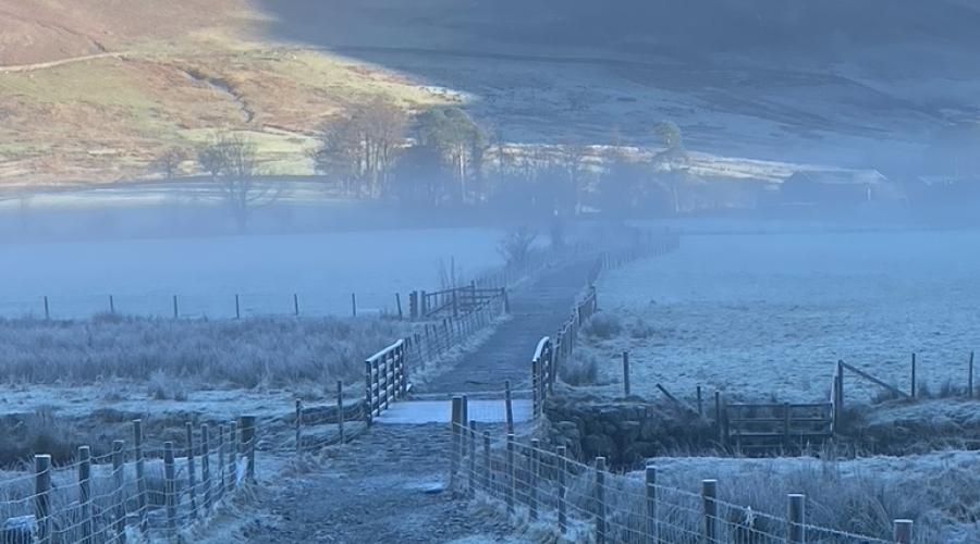 A path with fences either side leading through the misty frost-covered bottom of Buttermere Valley, with sun-covered green mountains rising in the background