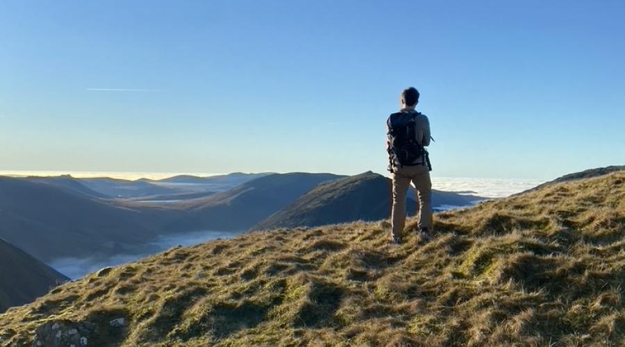 A walker standing with his back to the camera, standing on the grassy knolls of a hilltop, looking out across the mountains, with brilliant blue sky in the top half of the picture.