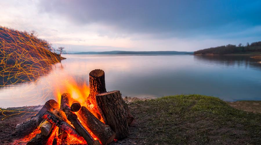 A campfire made of ember logs on the shoreline of a still blue lake in the twilight. Embers are blown from the fire leftwards out of frame, tracing orange lines across the picture.