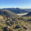 A view from the summit of Pillar looking out over surrounding dark green hilltops. Rocks and grass lie in the foreground, with cloud snaking through the valley between the mountains in the background and brilliant blue sky in the top third of the picture.