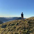 A walker standing with his back to the camera, standing on the grassy knolls of a hilltop, looking out across the mountains, with brilliant blue sky in the top half of the picture.