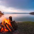 A campfire made of ember logs on the shoreline of a still blue lake in the twilight. Embers are blown from the fire leftwards out of frame, tracing orange lines across the picture.