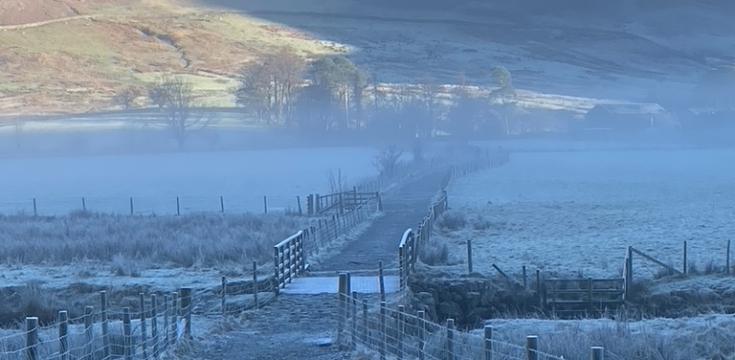 A path with fences either side leading through the misty frost-covered bottom of Buttermere Valley, with sun-covered green mountains rising in the background