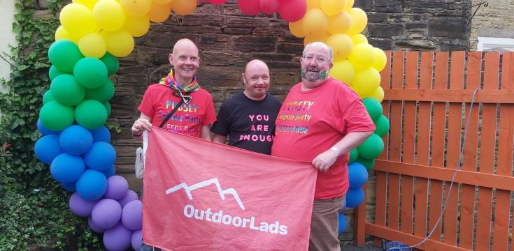 Three guys with Outdoorlads flag in front of a circular pride balloon display